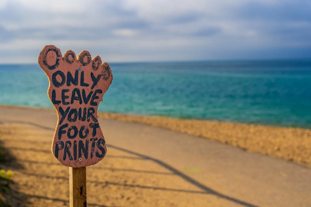 A footprint-shaped wooden sign on a beach path reading 'Only leave your footprints' with the sea in the background