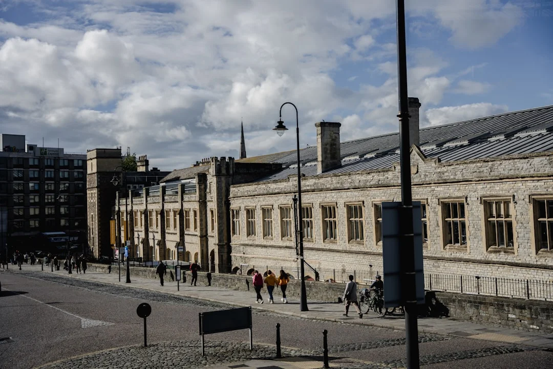 People walking on a sidewalk past a long, historic stone building in Bristol city centre under a cloudy sky