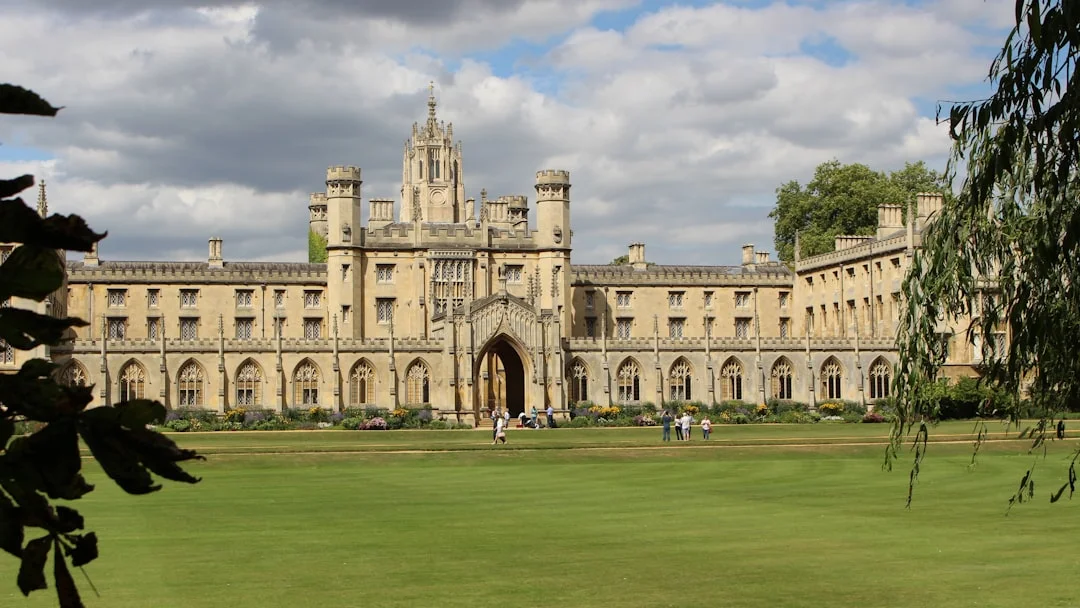 Historic stone college building with a central tower and arched gateway, fronted by a large green lawn