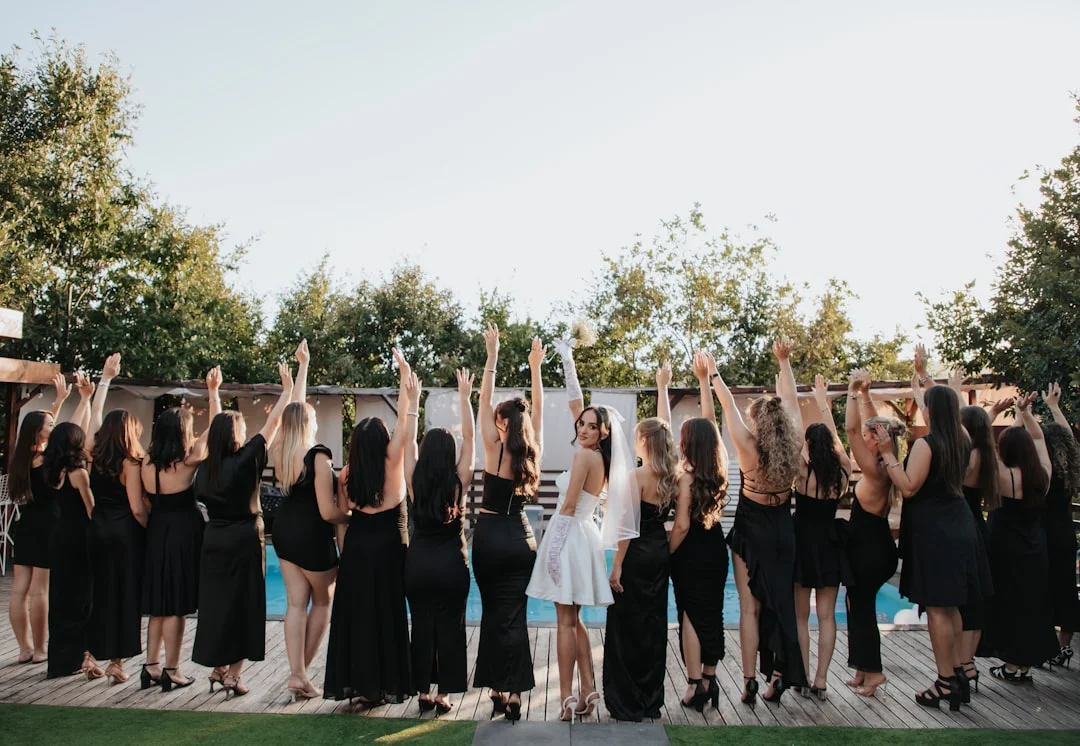 A bride in a white dress and veil stands with her bridesmaids in black dresses by a pool, arms raised