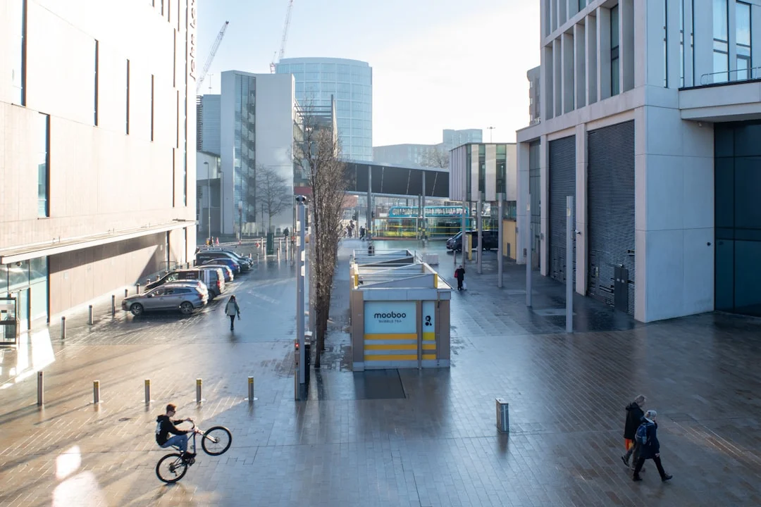 A cyclist performs a wheelie on a wide paved walkway between modern buildings in Liverpool