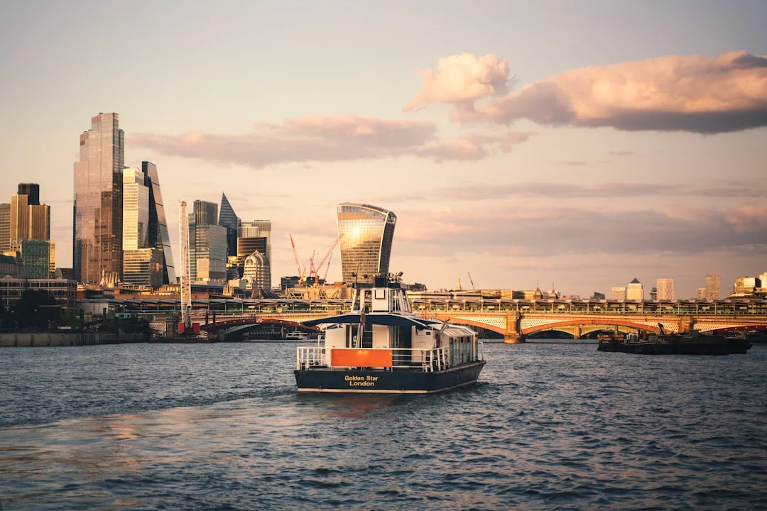 A boat cruises along the River Thames in London at sunset with the city skyline and a bridge in the background