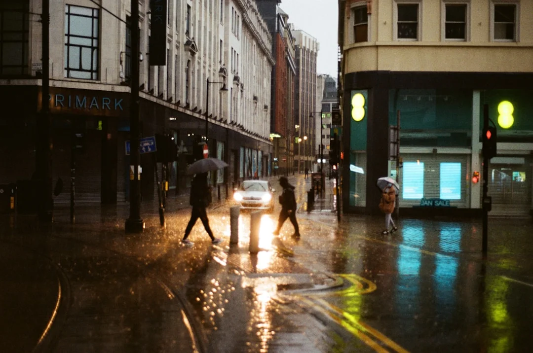 People with umbrellas cross a rainy Manchester street at night, with city lights reflecting on wet pavement