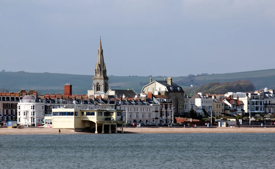 A view of Weymouth seafront from the water, featuring a sandy beach, historic buildings, and a tall church spire
