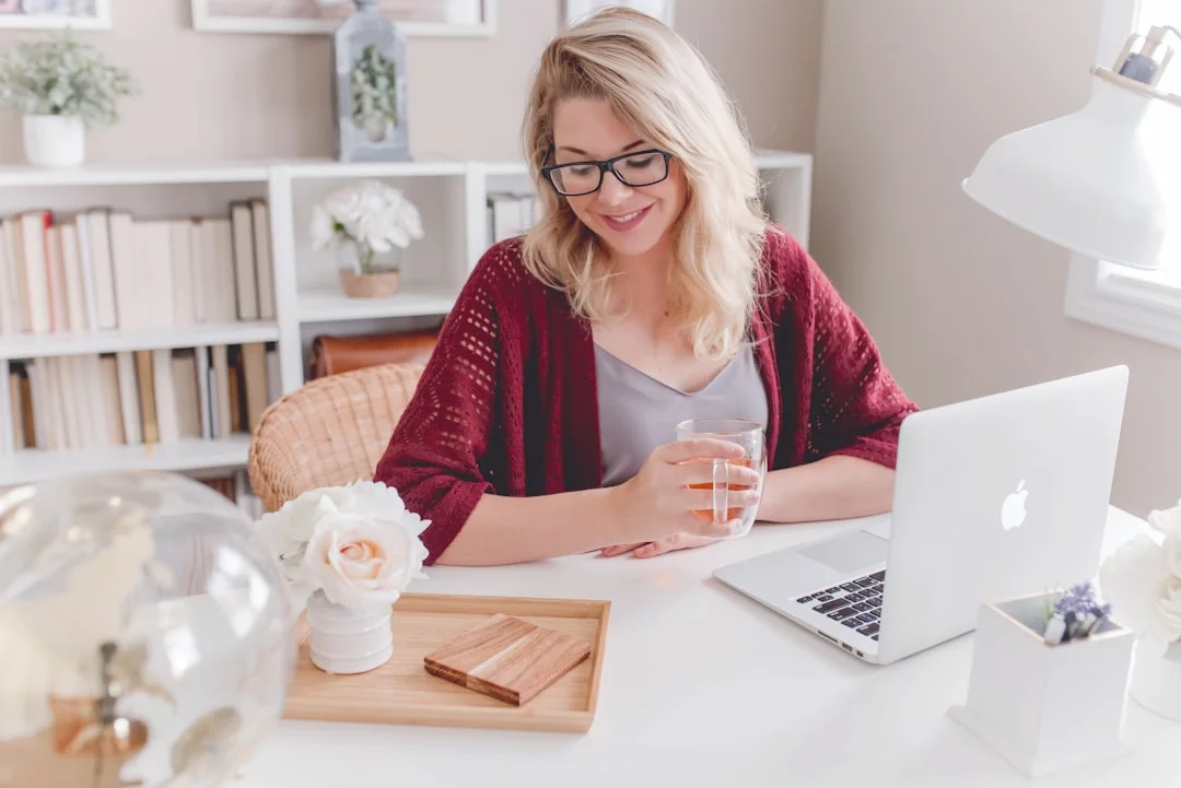 A blonde woman wearing glasses and a maroon cardigan sits at a desk with a laptop and a glass of water