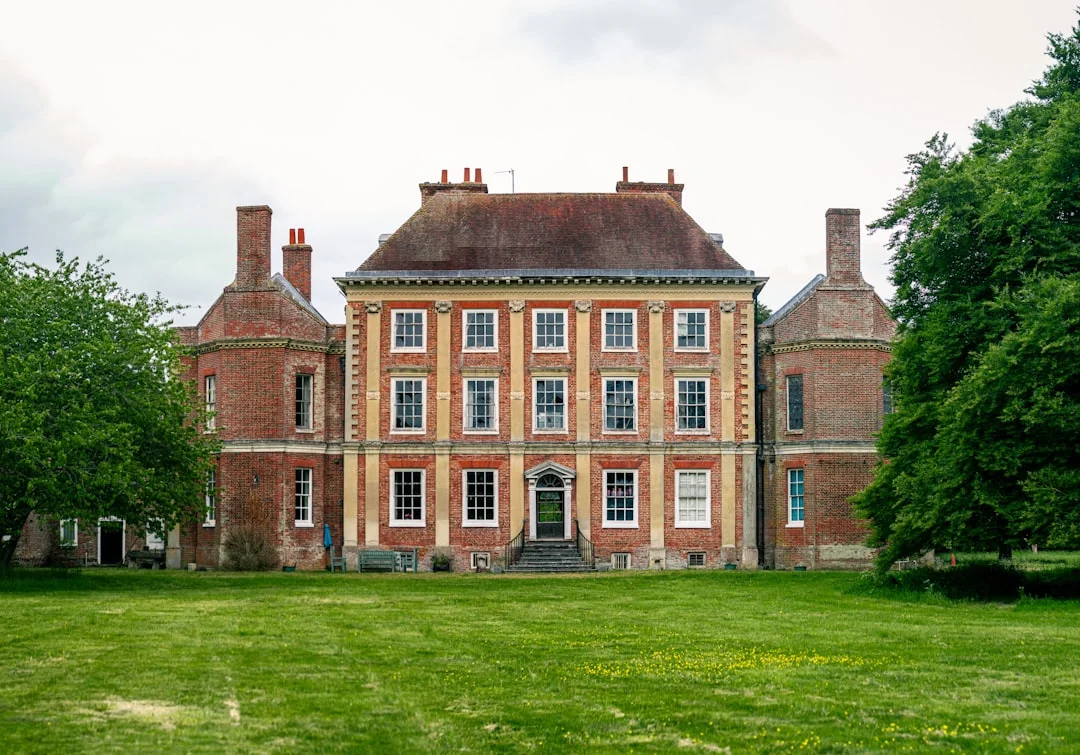 A grand red-brick manor house with symmetrical windows overlooking a large green lawn under a cloudy sky