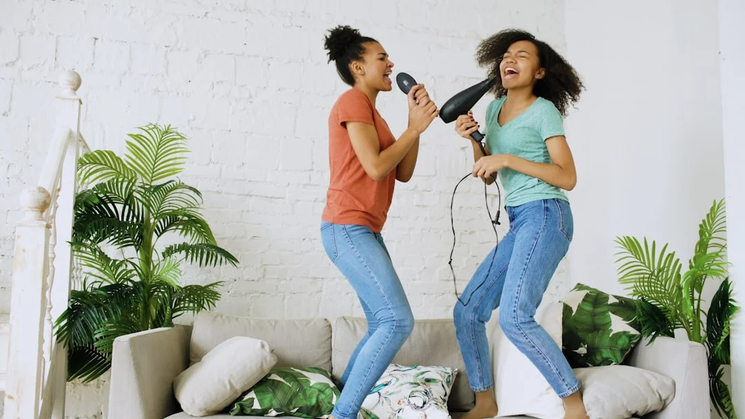 Two women jumping on a sofa, singing into a hairbrush and hairdryer in a bright living room