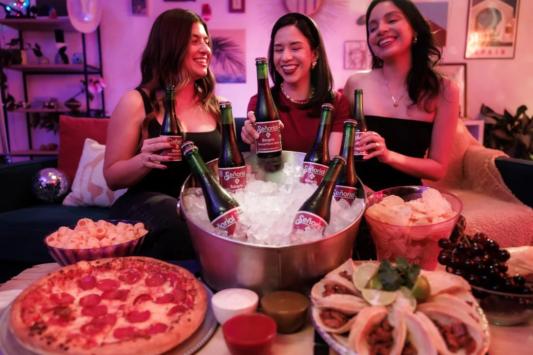 Three women smiling and holding beer bottles around a table with pizza, snacks, and an ice bucket of drinks