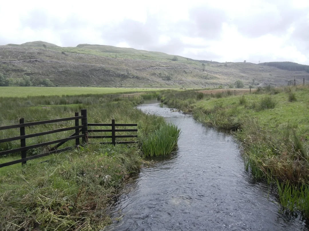 A small stream flows through green Scottish countryside with wooden fences and rolling hills under a cloudy sky