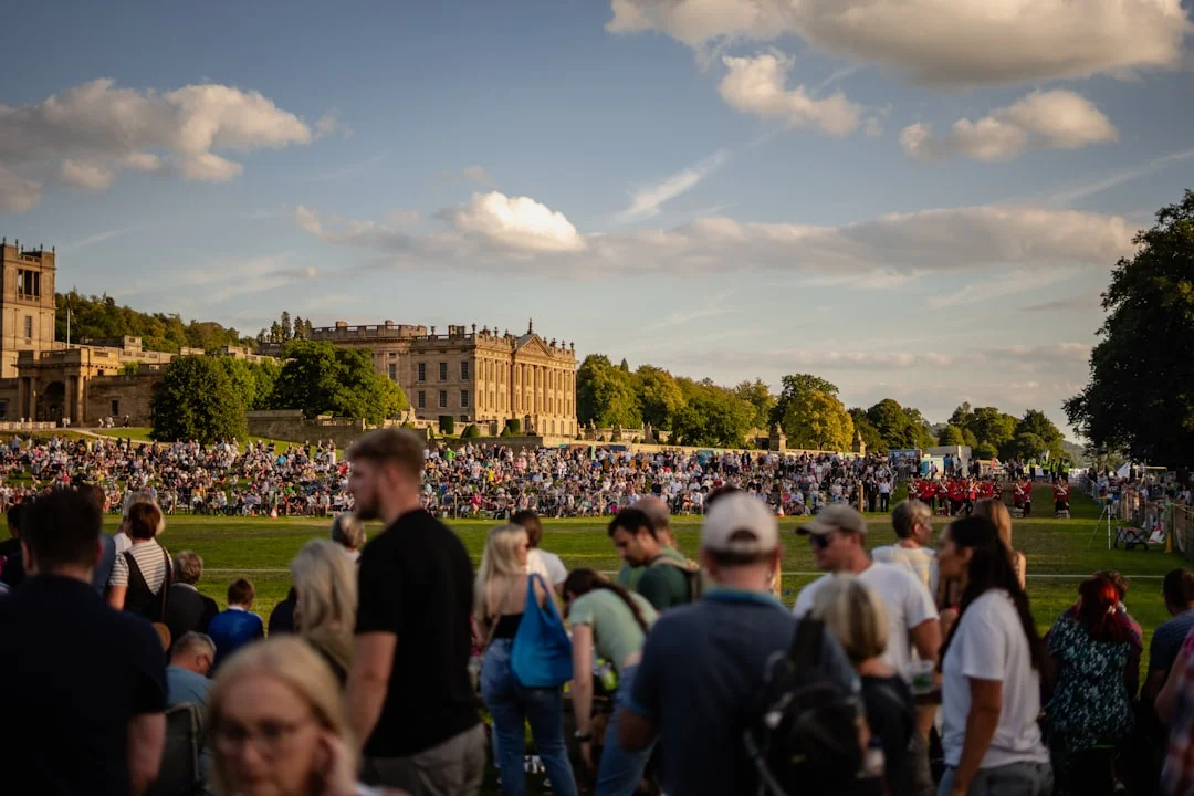 A large crowd of people gathered on a green lawn in front of a grand historic stately home under a blue sky