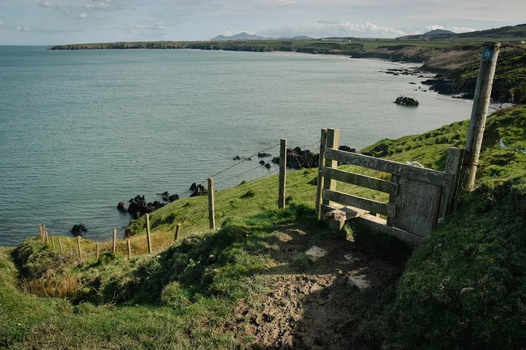 Wooden stile on a grassy cliff path overlooking a calm bay and distant headlands on the Llŷn Peninsula