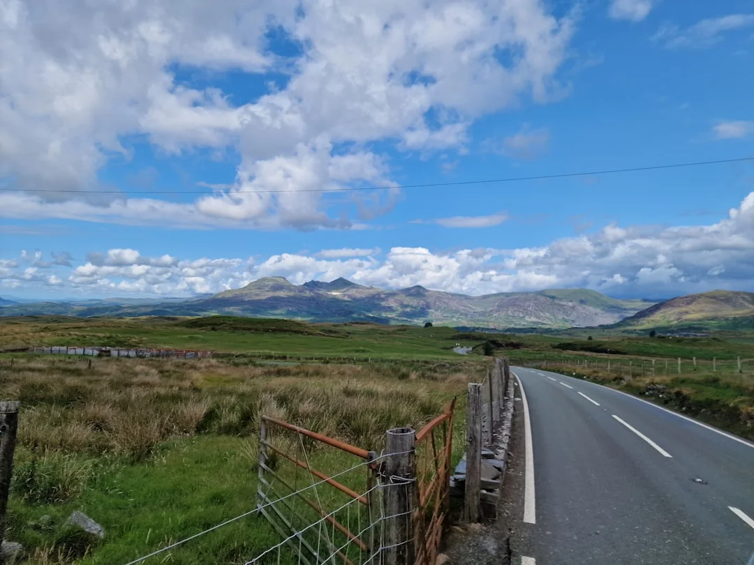 A winding road through green fields on the Llŷn Peninsula with mountains under a cloudy blue sky