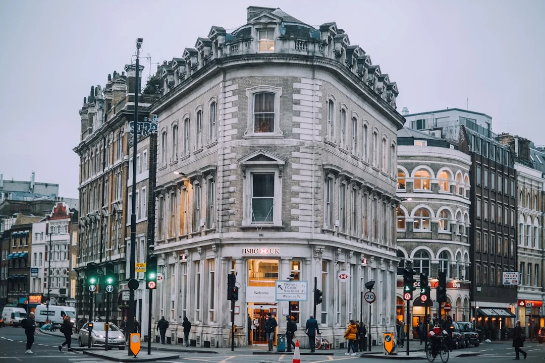 A grand stone corner building in London with large windows, street lights, and pedestrians at dusk