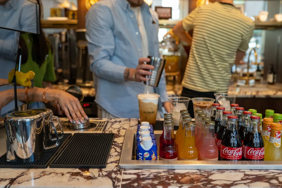 A person pours a drink from a shaker into a glass at a bar with various bottled beverages in the foreground