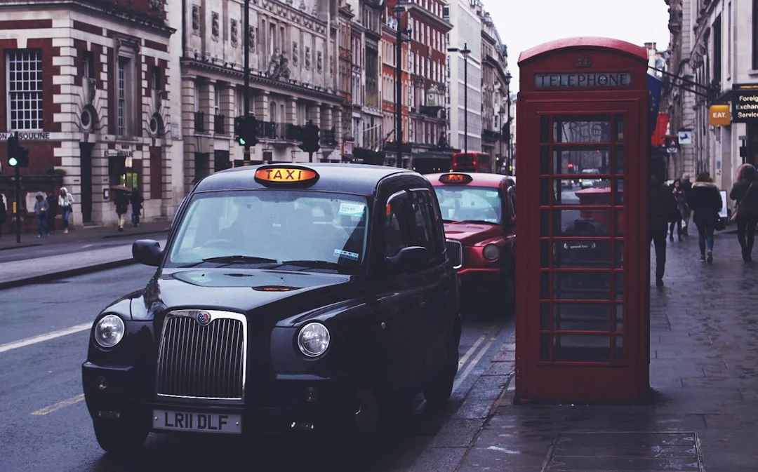 A black taxi cab parked on a London street next to a red telephone booth with pedestrians in the background