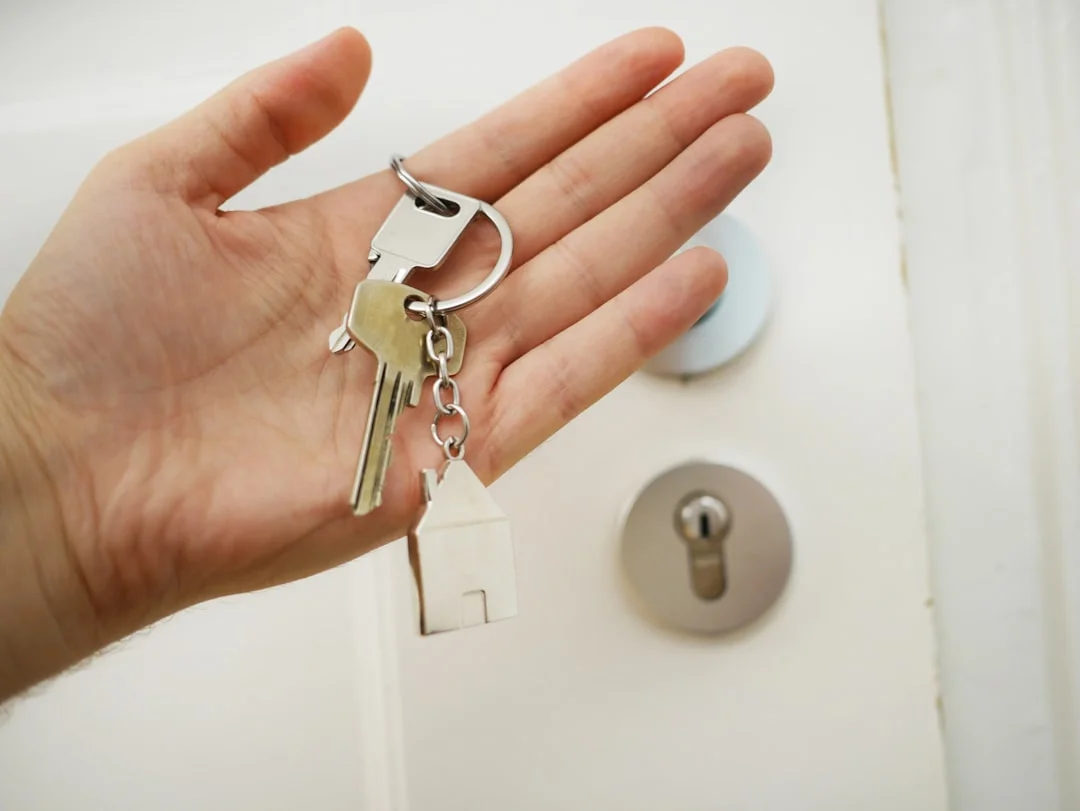 A hand holding a set of silver keys with a house-shaped keychain in front of a white door
