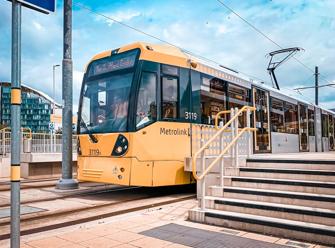 A yellow Manchester Metrolink tram stopped at a platform with stairs in the foreground under a blue sky