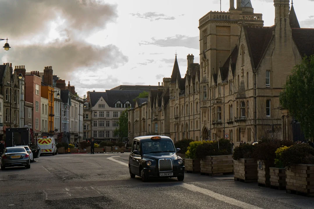 A black taxi drives down a street in Oxford past historic stone buildings under a cloudy sky