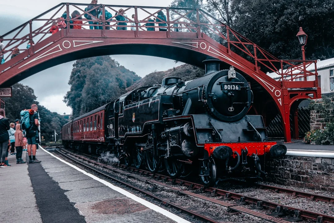 A vintage black steam engine pulling red carriages under a red footbridge at a station in Whitby