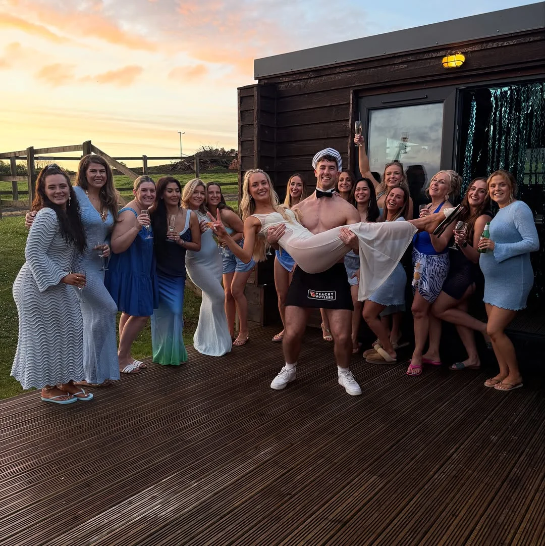 A butler in the buff serving drinks and hosting games for a laughing hen party group in their accommodation.