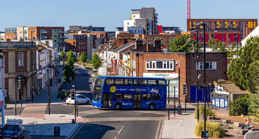 A blue double-decker bus drives through a town square with brick buildings and a clear blue sky