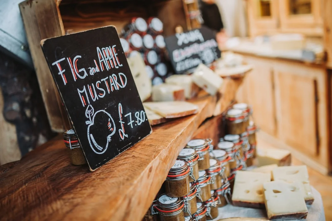 A wooden market stall with jars of fig and apple mustard, blocks of cheese, and a handwritten price sign