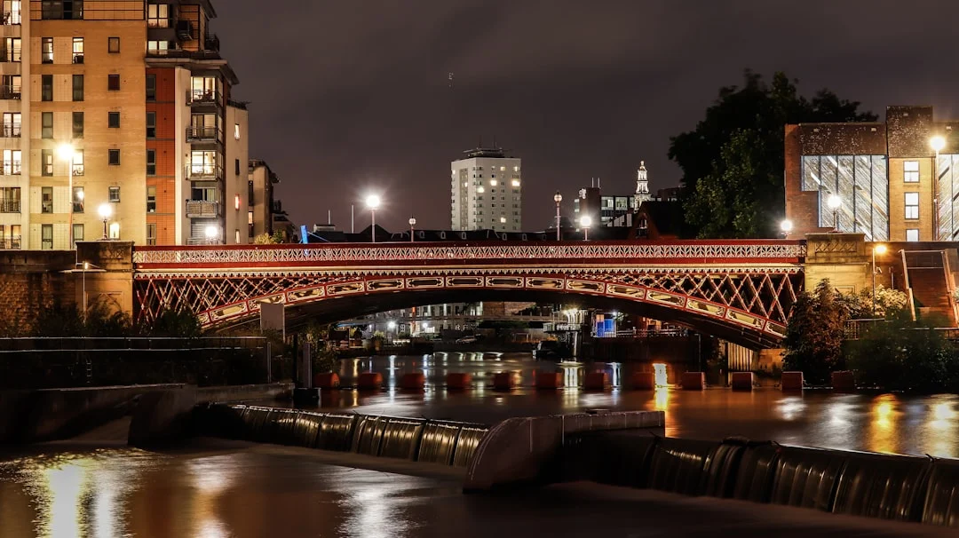 A lit-up bridge over the River Aire in Leeds at night with city buildings and a weir in the foreground