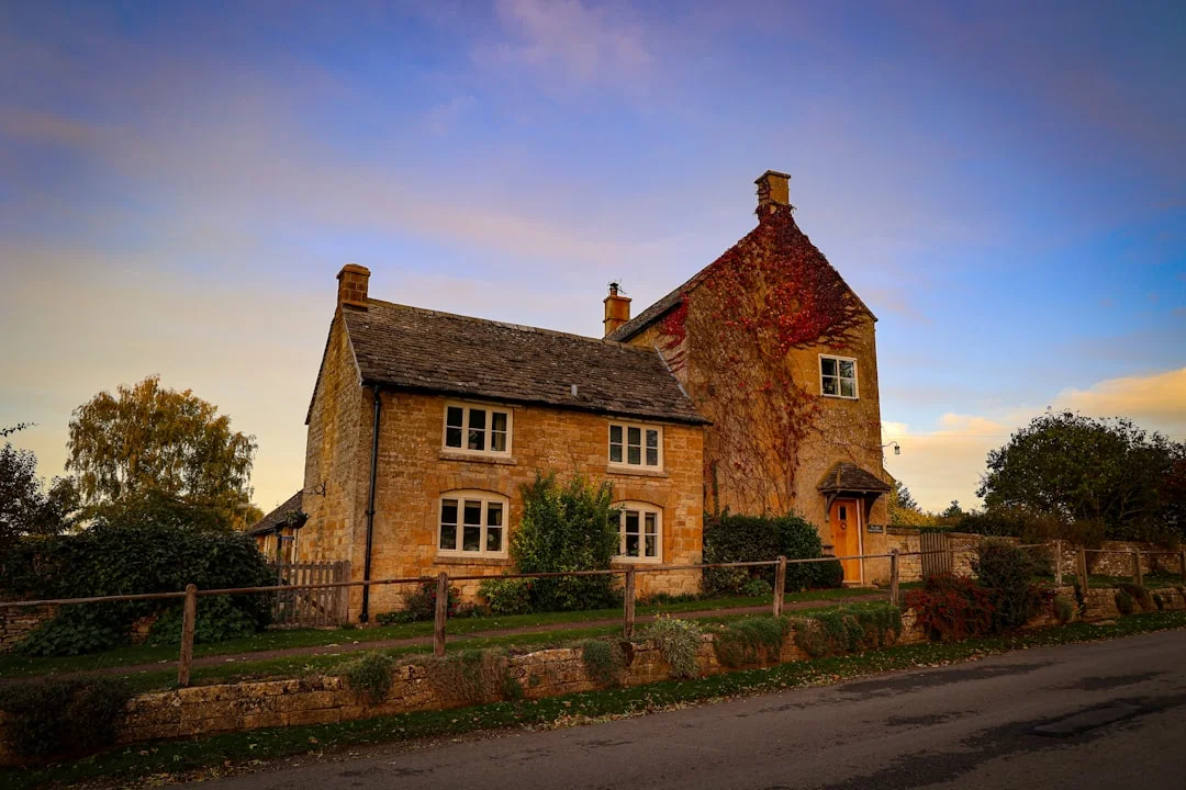 A traditional stone cottage with ivy-covered walls and a wooden fence under a golden sunset sky in Somerset