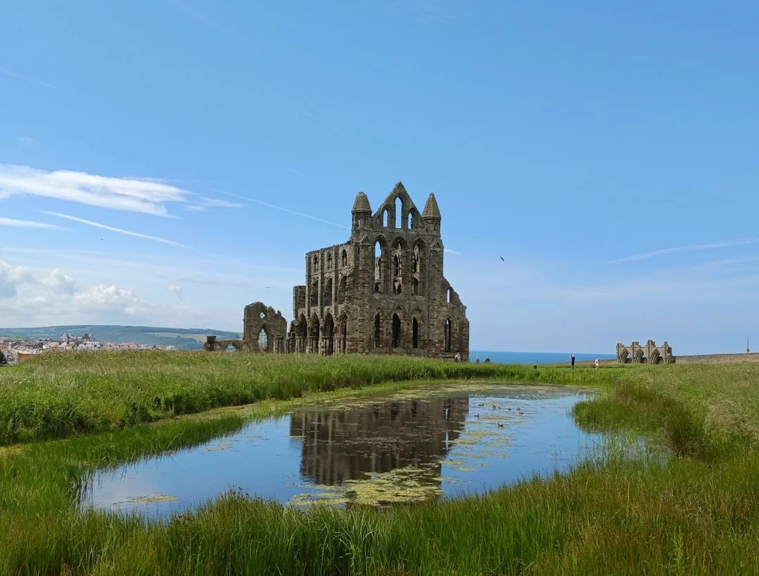 Ruins of Whitby Abbey standing on a grassy hill, reflected in a small pond under a clear blue sky