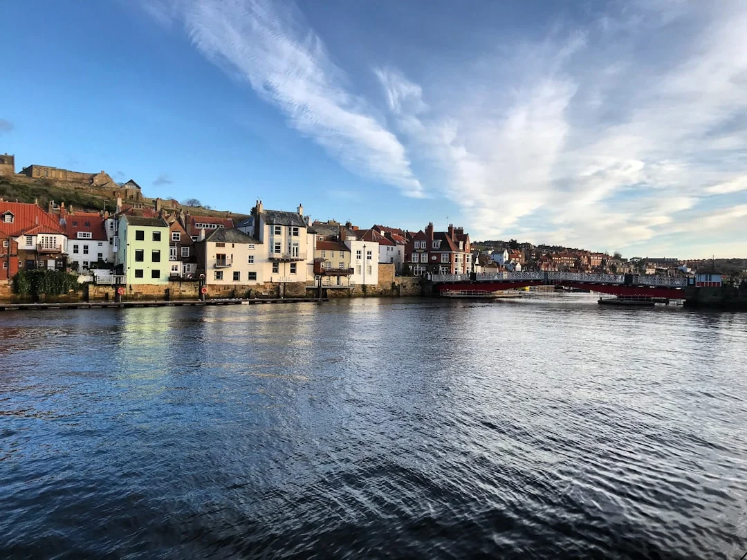 Colorful buildings line the Whitby harbor waterfront under a bright blue sky with wispy white clouds