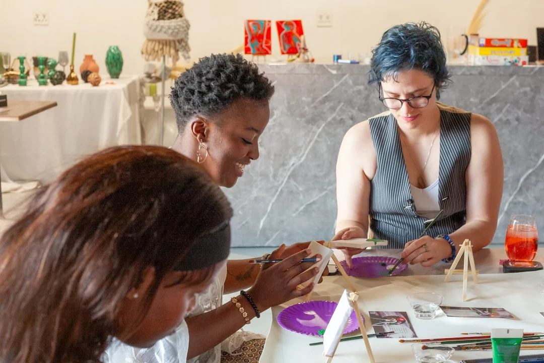 Three women smile while painting on small canvases and crafting at a table during a creative workshop