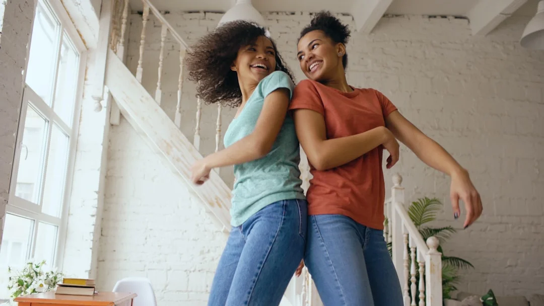 Two women dancing back-to-back and laughing in a light-filled room with a wooden staircase