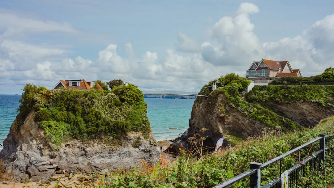 Two houses perched on rocky cliffs overlooking a turquoise sea under a cloudy sky in Newquay