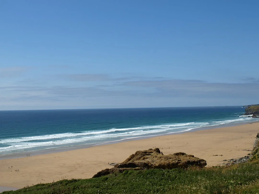A wide sandy beach with ocean waves, seen from a grassy cliff under a blue sky