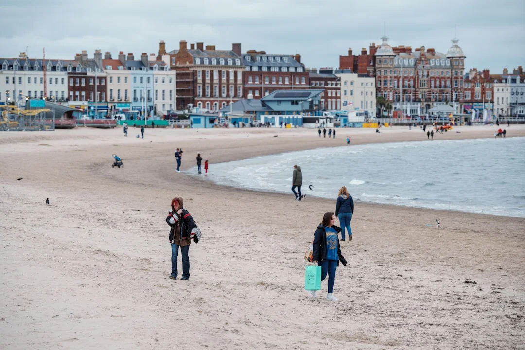 People walking on Weymouth beach with a backdrop of grand coastal buildings under a cloudy sky