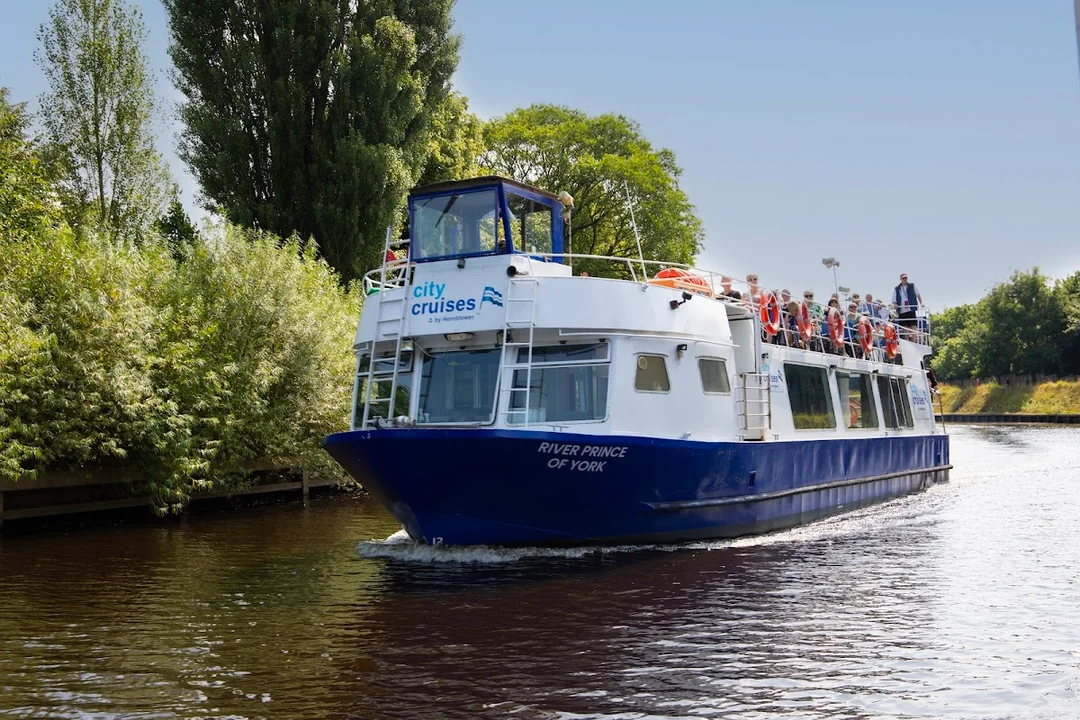 A tour boat cruising down the River Ouse in York on a sunny day, with landmarks in the background.